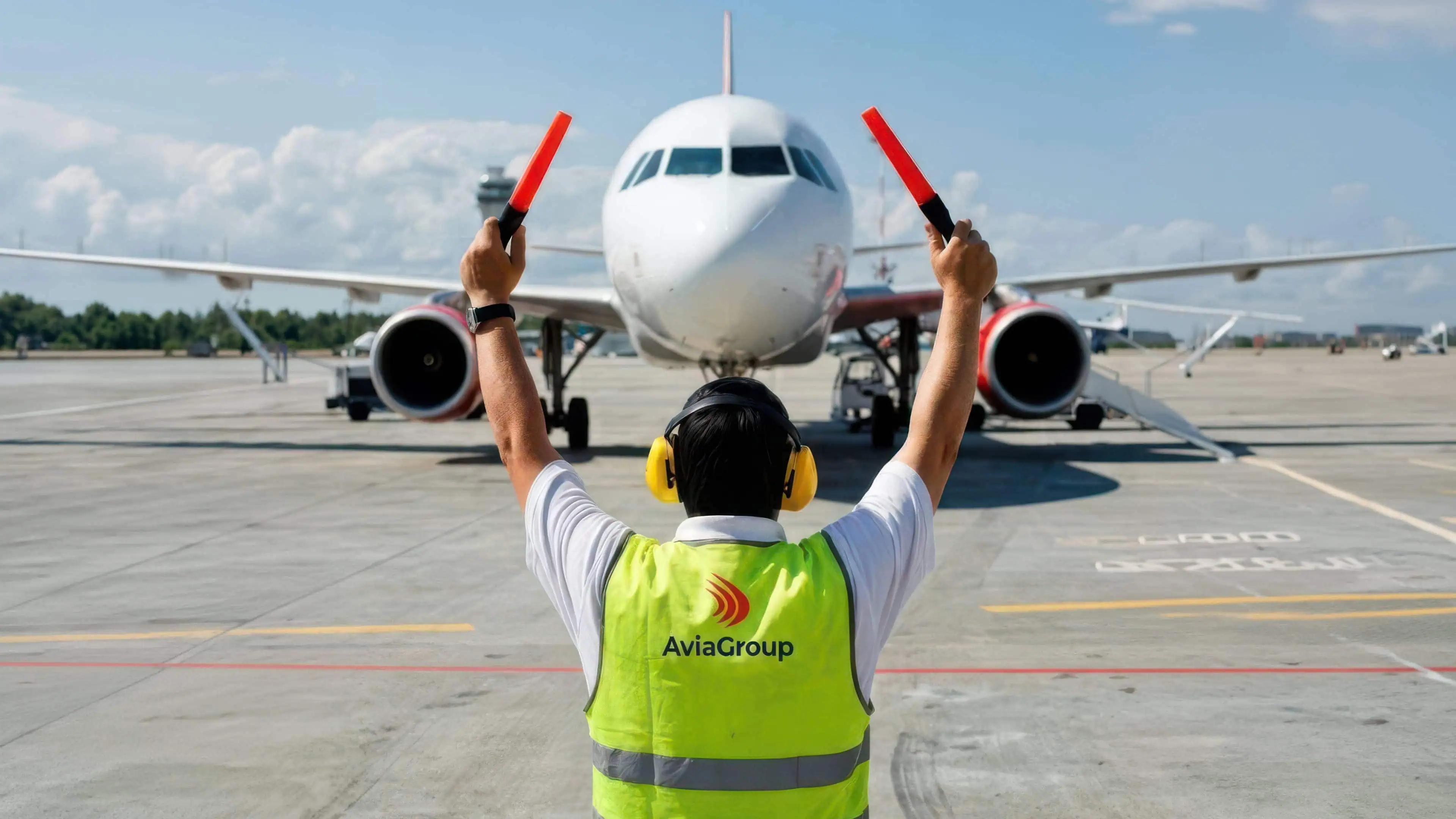 AviaGroup marshaller in a hi-viz vest raising red wands to guide an aircraft on the apron